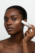 A cosmetic stick product with its cap removed, standing upright on a clean white studio background. The product has a taupe-beige label, a white cap, and a light beige cream stick visible at the top.